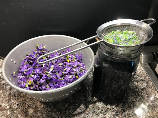 fresh violet flowers in a colander shown next to the same amount of flowers that have been steeped in boiling water, then pressed and squeezed to remove as much liquid as possible